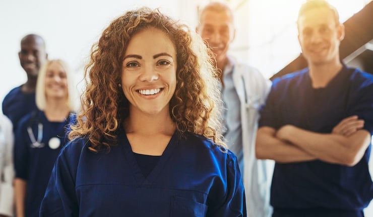 A doctor smiling at the camera while working at a hospital with other healthcare professionals in the background.